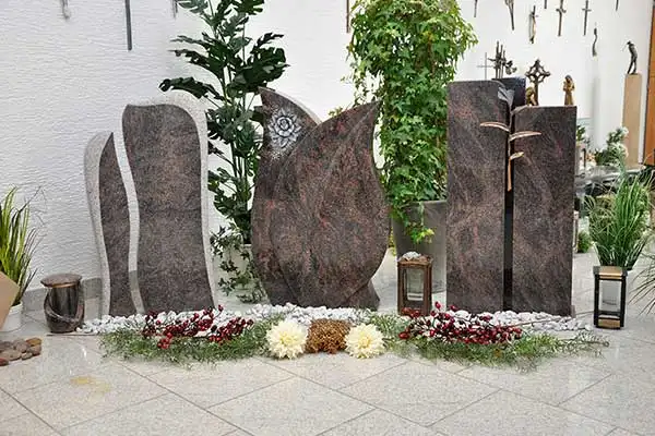 Rotbraune Grabsteine mit Rosenornament und metallischem Schmuck zwischen Pflanzen
