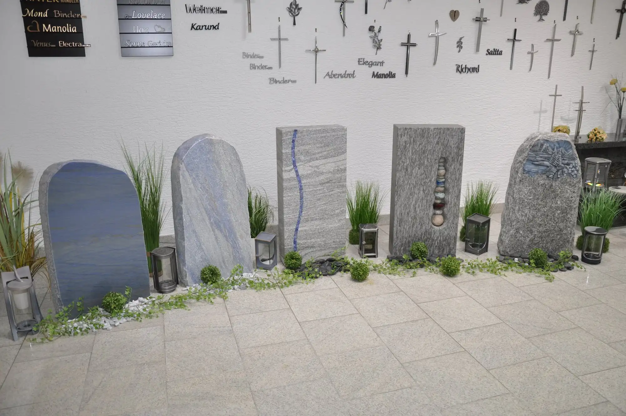 Grabsteine in Blau- und Grautönen, einer mit blauem Wellenband, einer mit Kieseln, rechts ein rauer Findling mit Relief