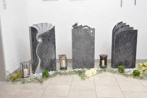 Drei dunkle Grabsteine mit Wellenrelief, Vogelfigur und Buchform
